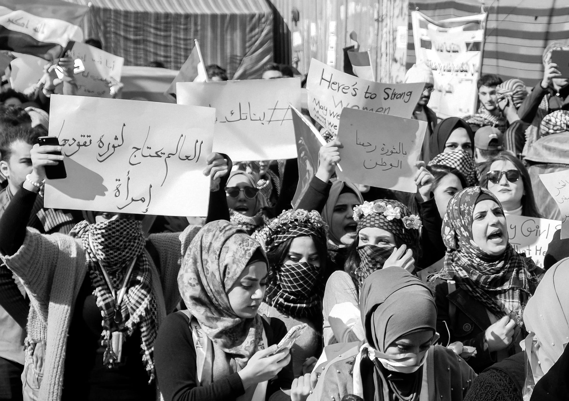 Women march at a protest in Iraq holding Arabic and English signs, one reading 'Here's to strong women'