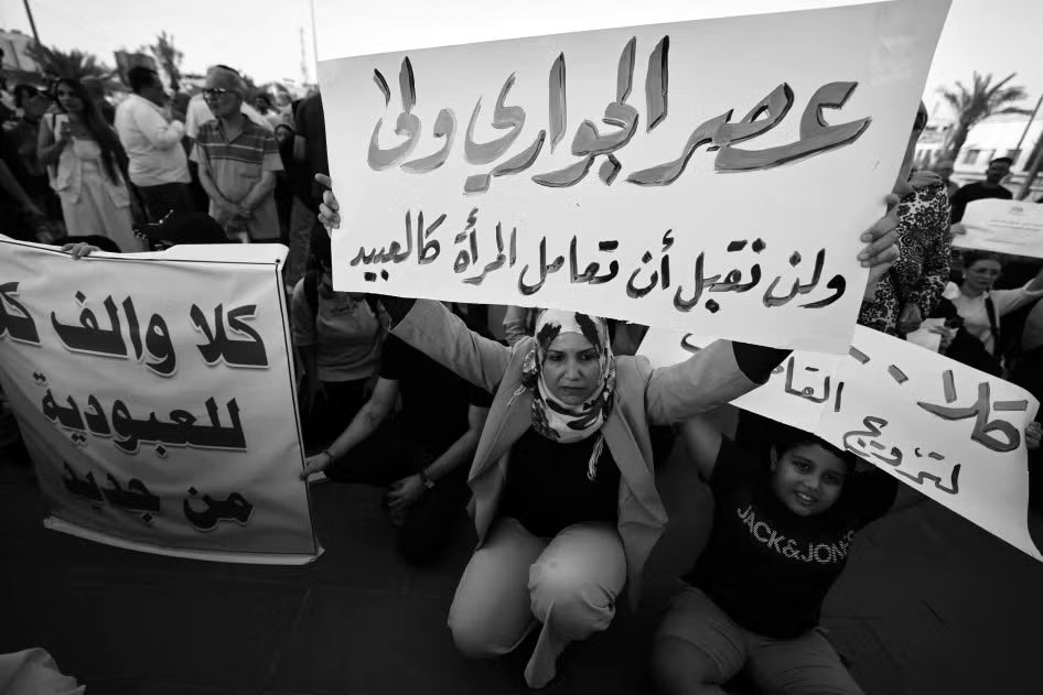 A woman sits in a crowd and holds up an Arabic sign saying women will not be treated as slaves