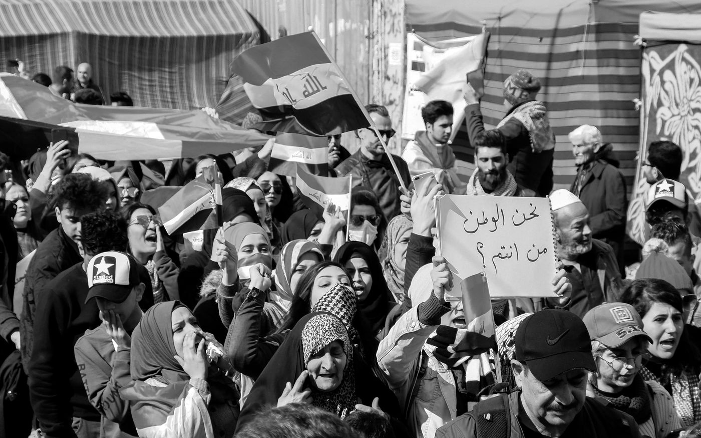 A woman holds a handwritten sign in Arabic at a crowded march in Baghdad, among Iraqi flags