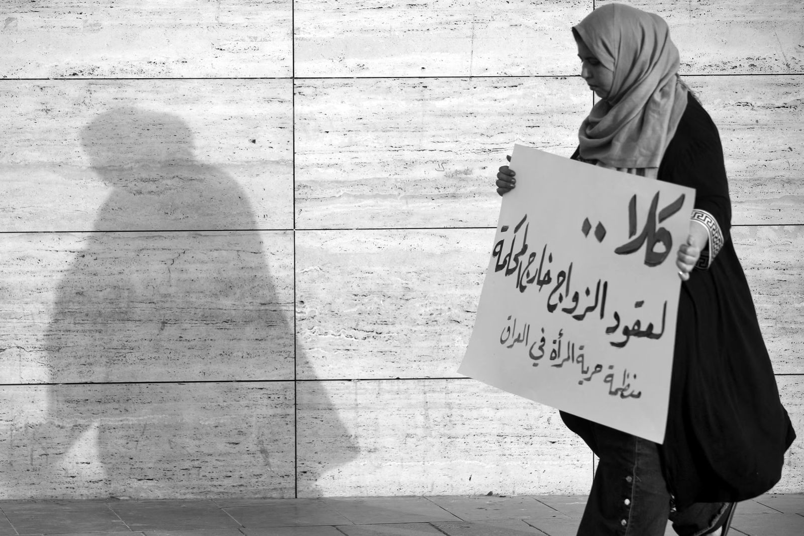A woman in a headscarf walks past a stone wall holding an Arabic placard, her shadow on the wall beside her